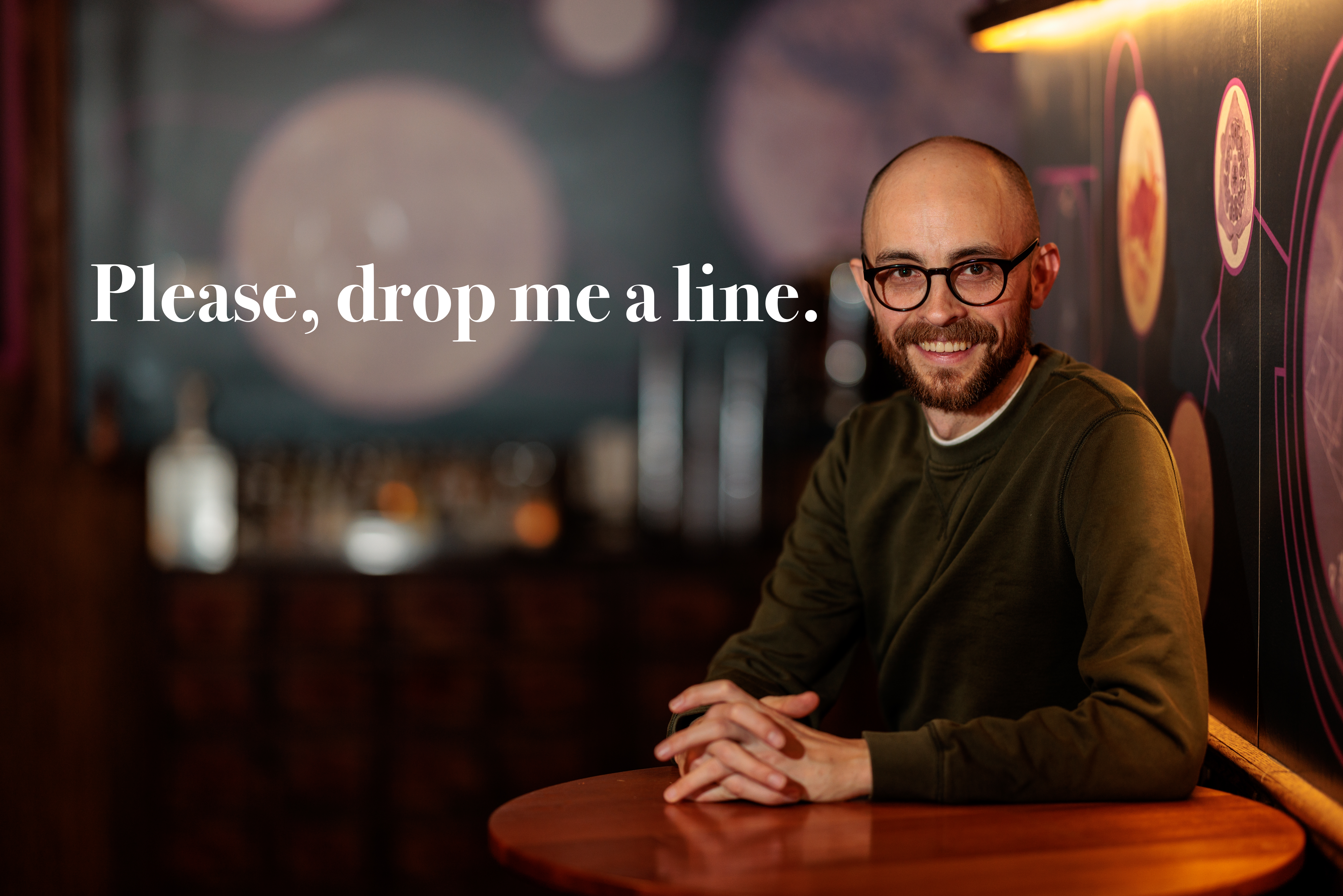 a strange, bald man with glasses, nearly forty years old, sits at a hightop table in a comedy club, beckoning you to contact him. in white text to the left of him, it reads, 'Please, drop me a line.' he begs, like a pathetic loser.